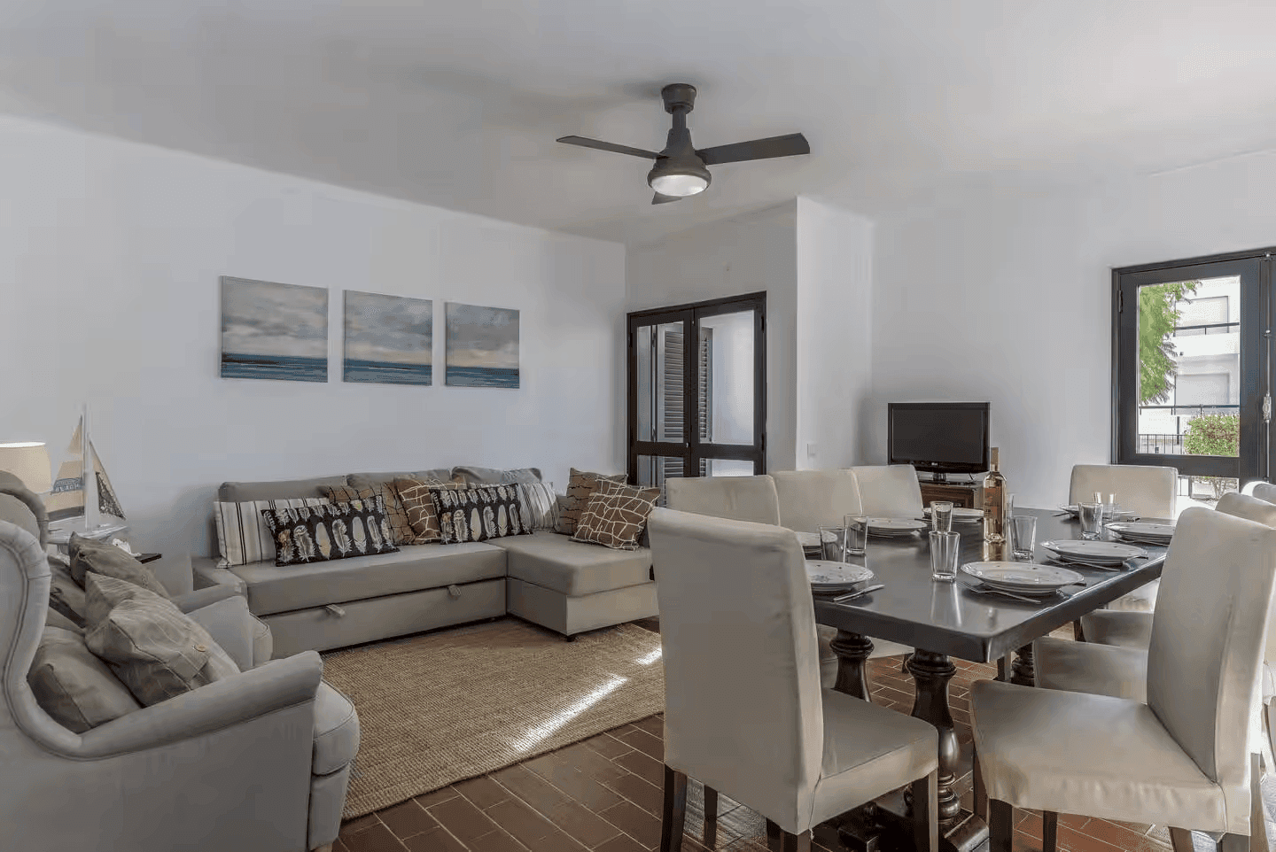 Sala de estar e jantar com sofá cinza, mesa posta e quadros de paisagens marinhas.